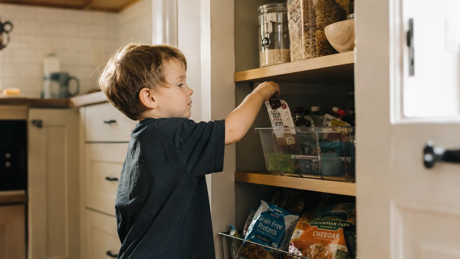 Child reaching into a clear snack bin on a low pantry shelf, self-serve snack station setup
