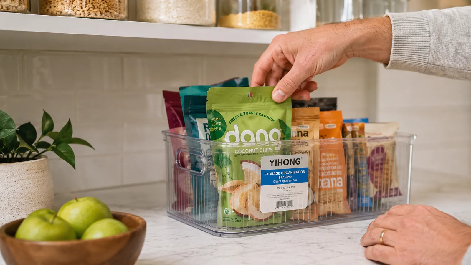 Hand reaching into a clear pantry bin to grab a snack pouch, snacks visible through the bin wall